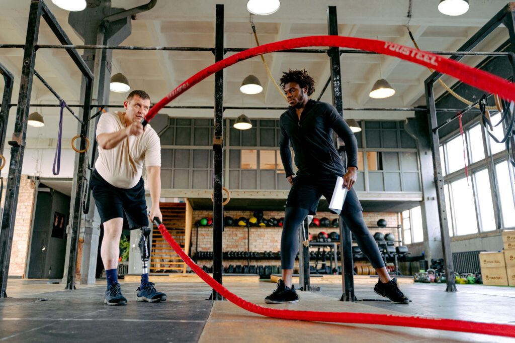 An adult male with a prosthetic leg exercises with ropes, guided by a personal trainer in a gym.