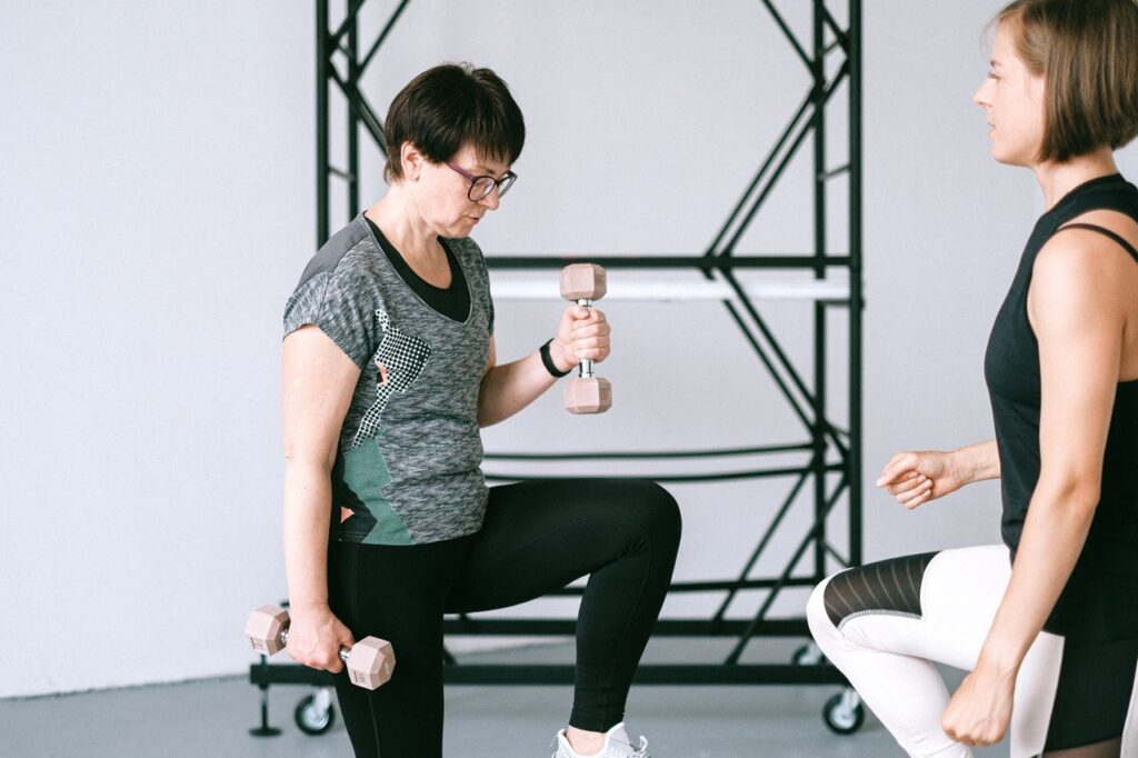 Elderly woman lifting dumbbells with guidance from a trainer indoors.